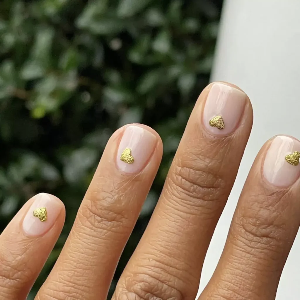 Image of a clear or pale pink nail base featuring a tiny gold heart sticker near the cuticle of each nail. by themimid