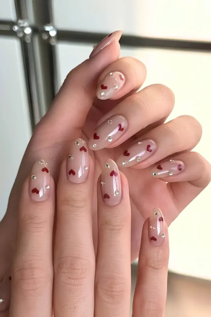 Nude manicure stacked with tiny gems and simple red heart decals. by 42nail.studio